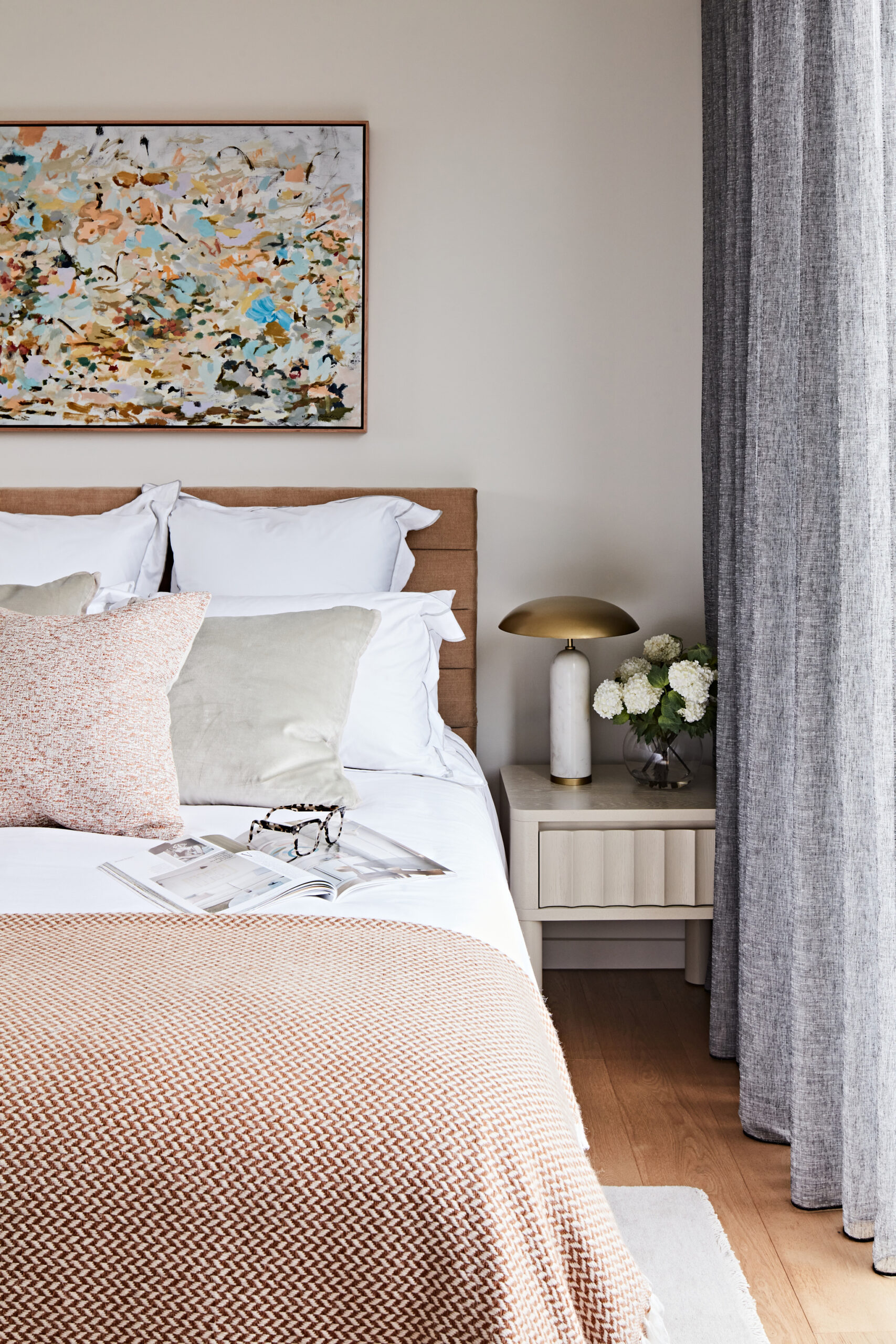 Modern Australian bedroom featuring layered linen bedding, textured soft furnishings, abstract artwork and sheer charcoal curtains in a Hampton downsizer apartment.