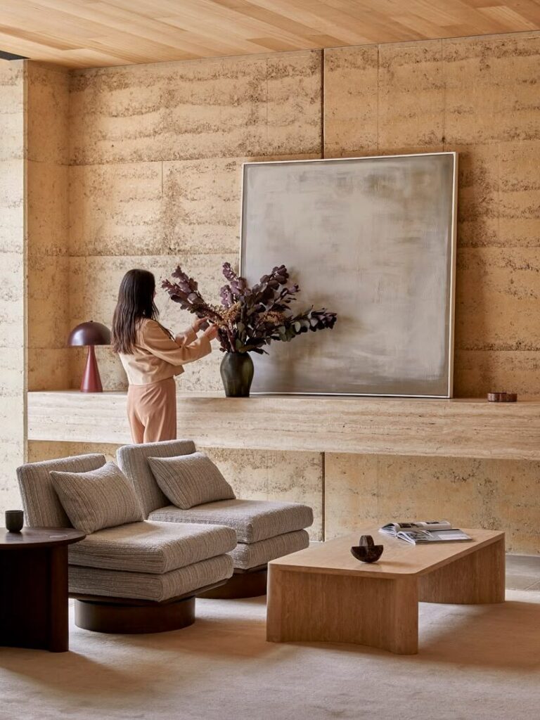Contemporary living room featuring natural stone walls, sculptural upholstered lounge chairs, timber coffee table, oversized abstract artwork and warm neutral furnishings.